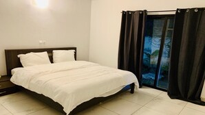 Una cama bien hecha con ropa de cama blanca en una habitación con suelo de baldosas y una ventana con cortinas oscuras.