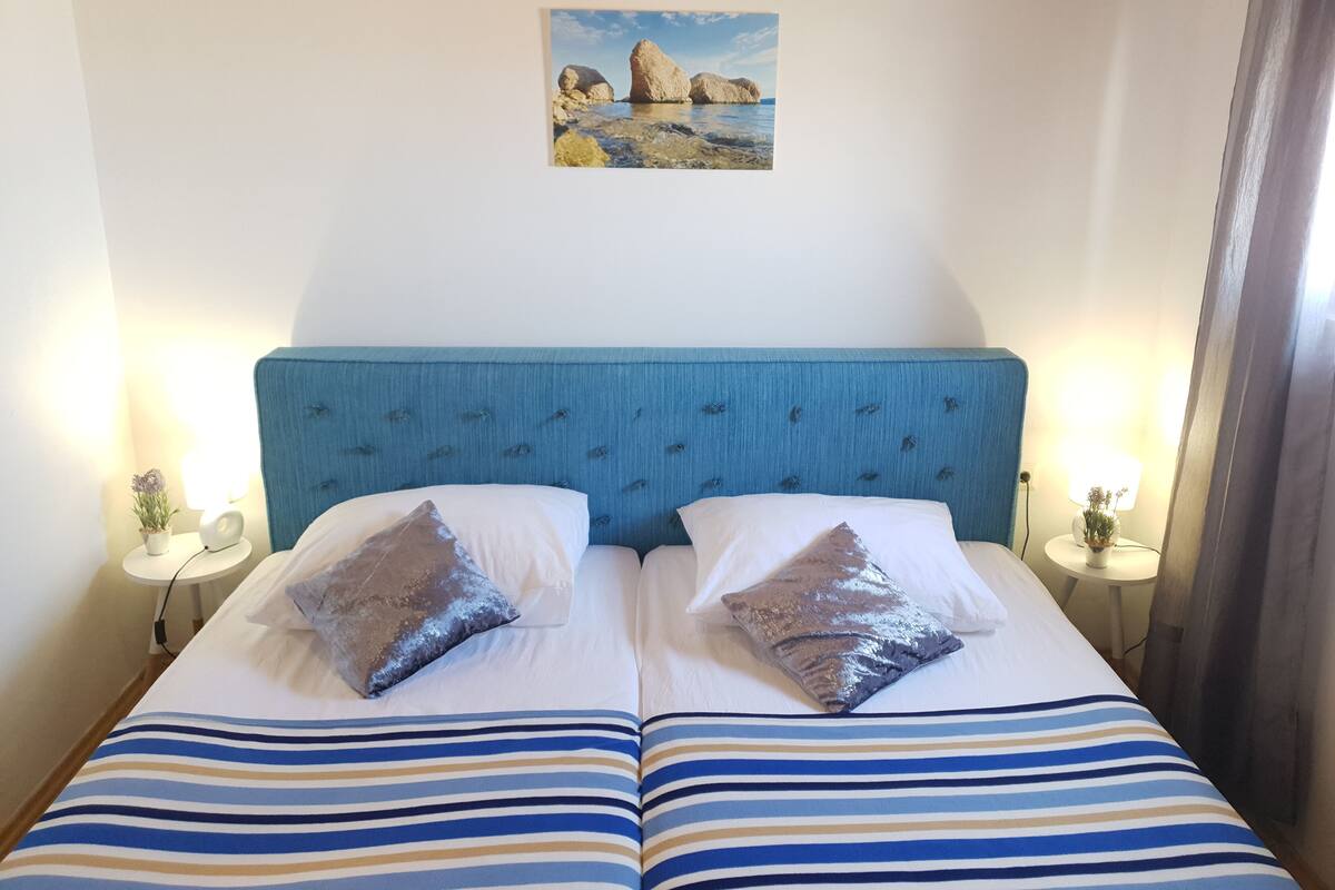 Una cama con cabecera azul, dos almohadas y ropa de cama a rayas. Encima de la cama, hay un pequeño cuadro en la pared.
