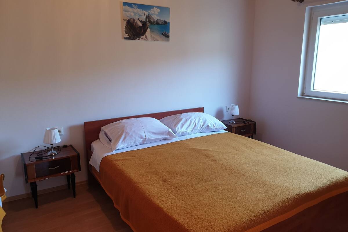 Un dormitorio con cama, mesita de noche, lámpara, ventana y un cuadro en la pared.