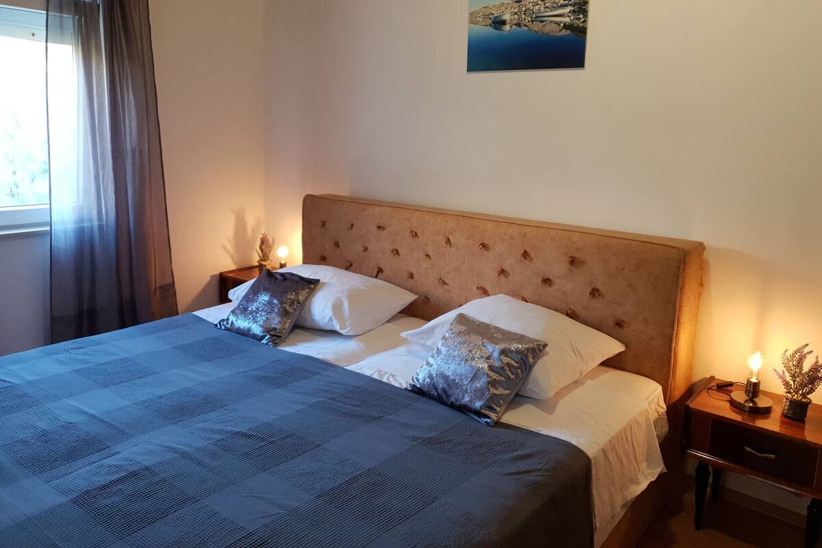 Una cama bien hecha con una colcha a cuadros, dos almohadas y un cabecero. Hay una mesita de noche con una lámpara y una pequeña planta en maceta. Un cuadro de una escena costera cuelga en la pared.