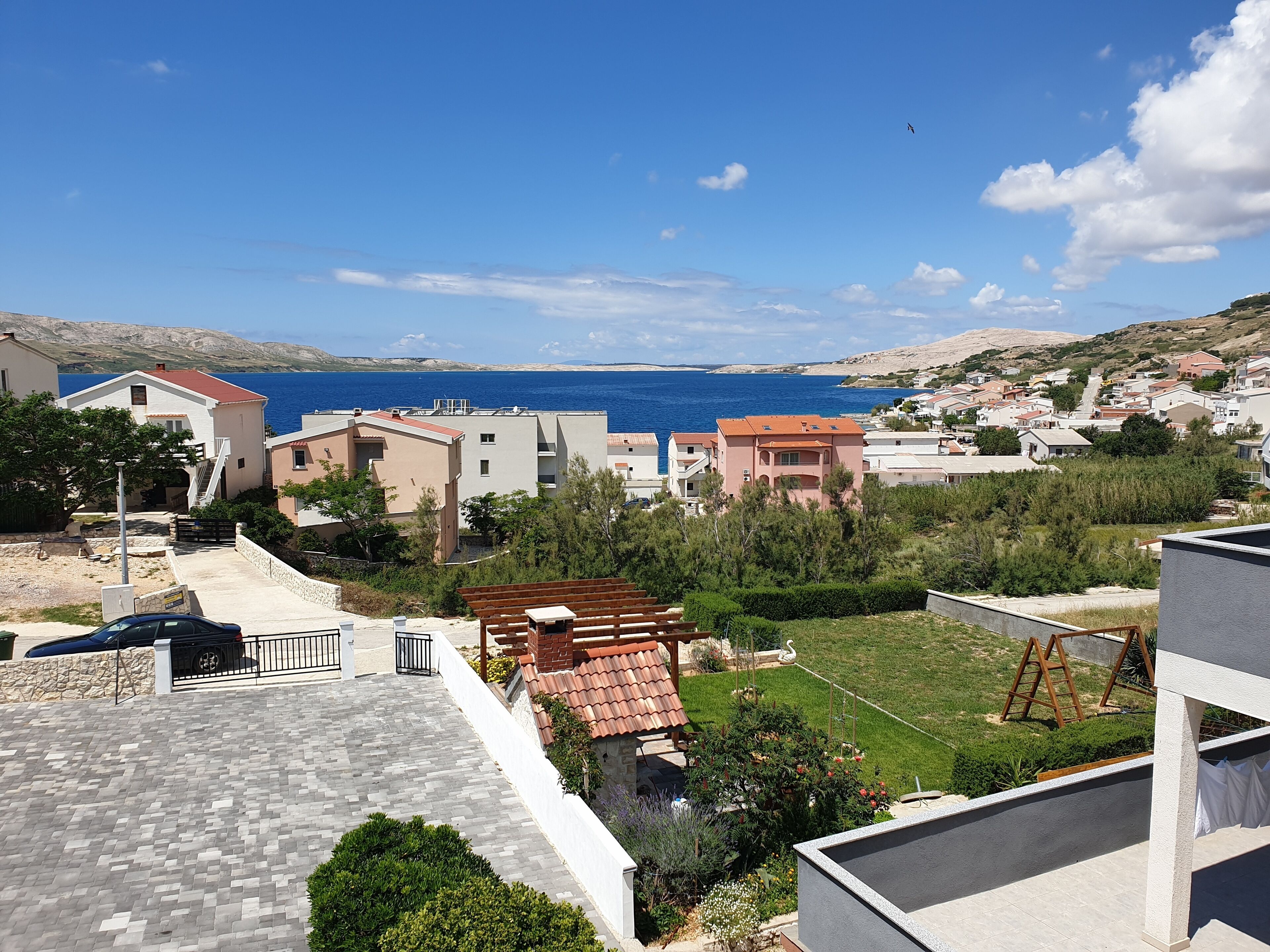 Zona residencial con vista al mar, con casas de techos rojos y blancos, un área pavimentada y un césped.