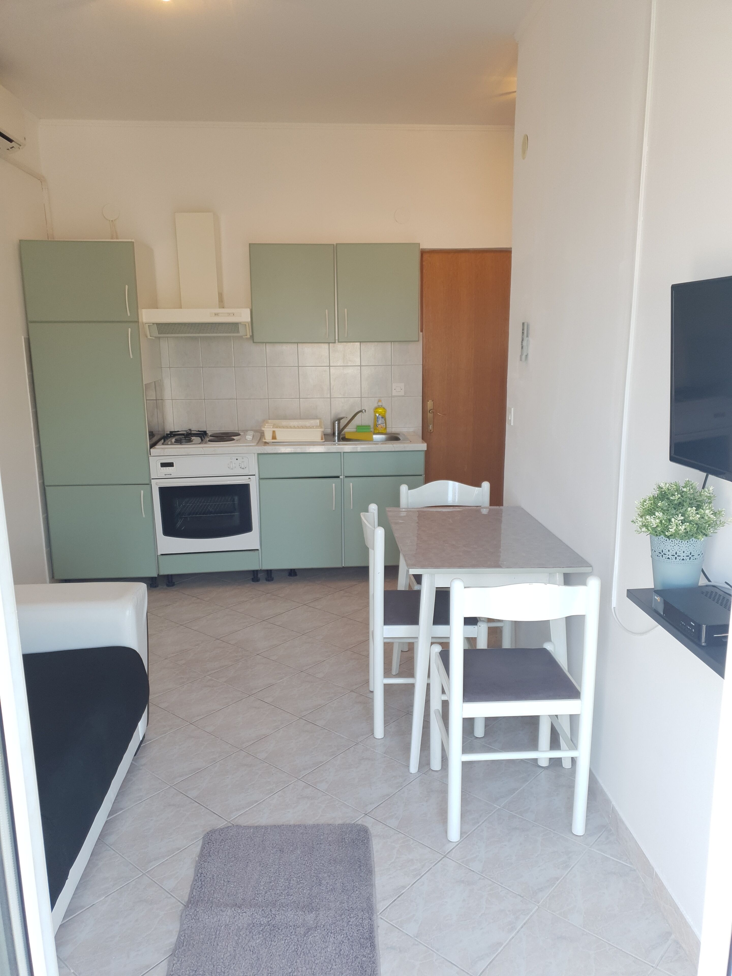 A compact kitchen with green cabinets, a stove, and a sink. There is a dining table with chairs and a small rug on the floor.
