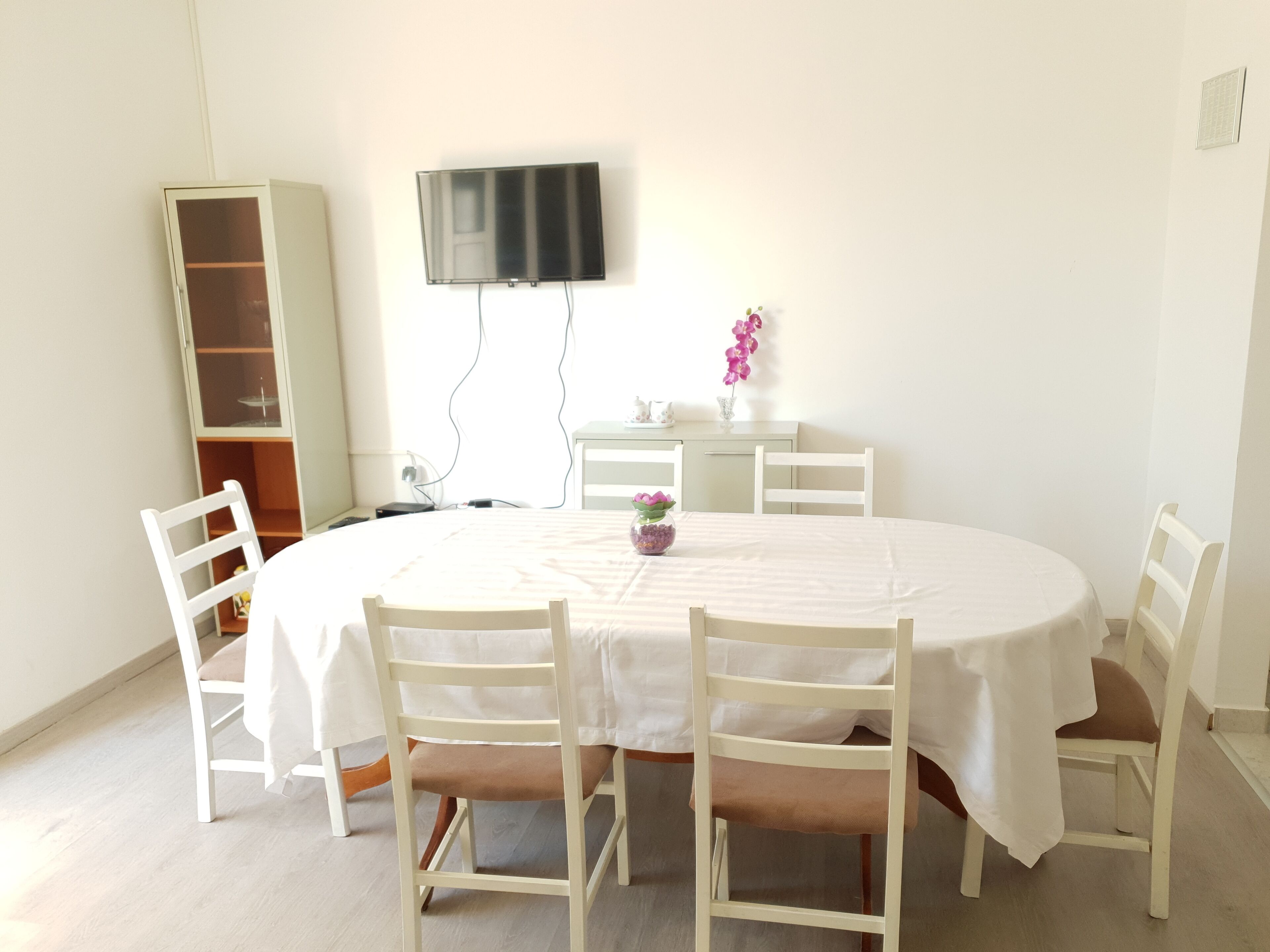 Un comedor con una mesa redonda, sillas blancas, un televisor montado en la pared y un mueble bajo.