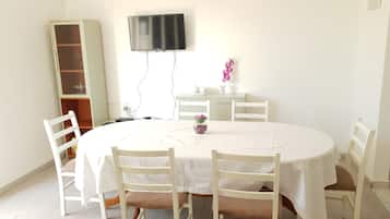 Un comedor con una mesa redonda, sillas blancas, un televisor montado en la pared y un mueble bajo.