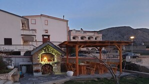 A stone-built outdoor fireplace with a fire, surrounded by wooden benches and a pergola, set against a hillside backdrop. - Apartments Dalmatinka Metajna Novalja (Novalja)