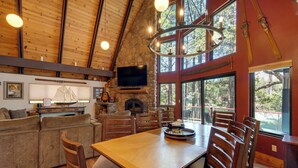 Interior acogedor de una cabaña con chimenea de piedra, mesa de comedor de madera y ventanas amplias con vistas al bosque.