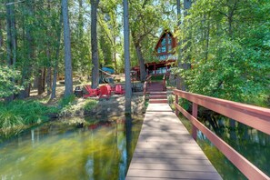 A wooden bridge leading to a cabin with a red roof, surrounded by trees and a calm body of water.