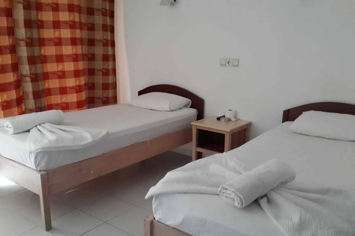 A hotel room with two single beds, each with a neatly folded towel, a wooden nightstand with a lamp, and a red and beige checkered curtain.
