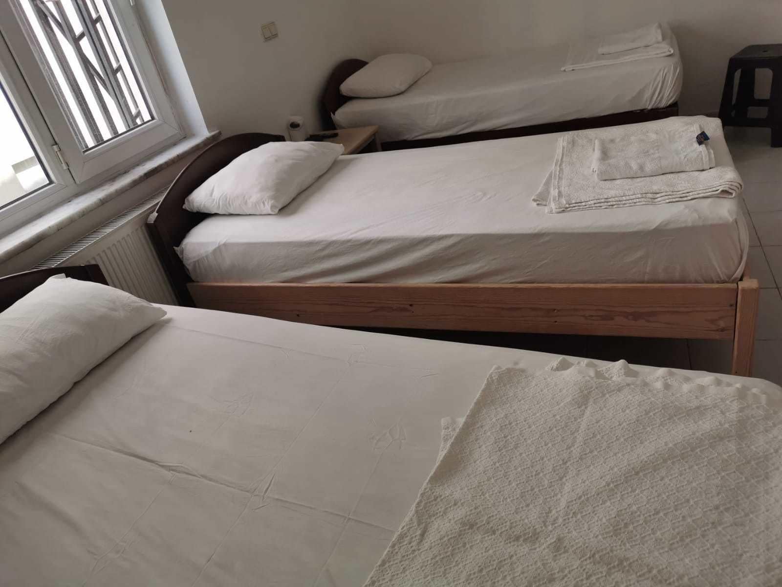 A room with two single beds, each with a wooden frame and white bedding. There is a window on the left side, and a radiator is visible on the wall.