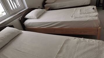 A room with two single beds, each with a wooden frame and white bedding. There is a window on the left side, and a radiator is visible on the wall.