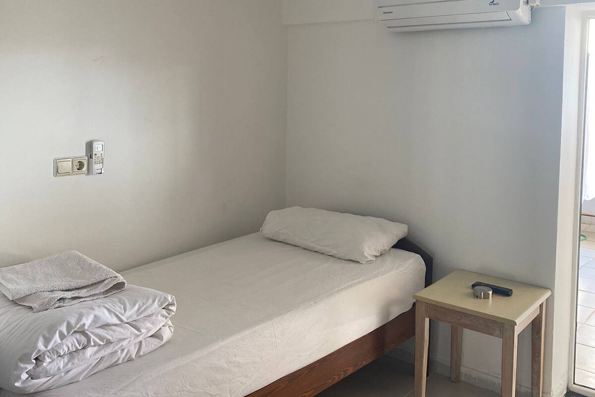 A small, single-bed room with a wooden frame, a bedside table, a wall-mounted air conditioner, and a window.