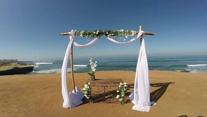 Bryllupsopsætning på stranden med en træbue, hvide draperinger og blomsterpynt.