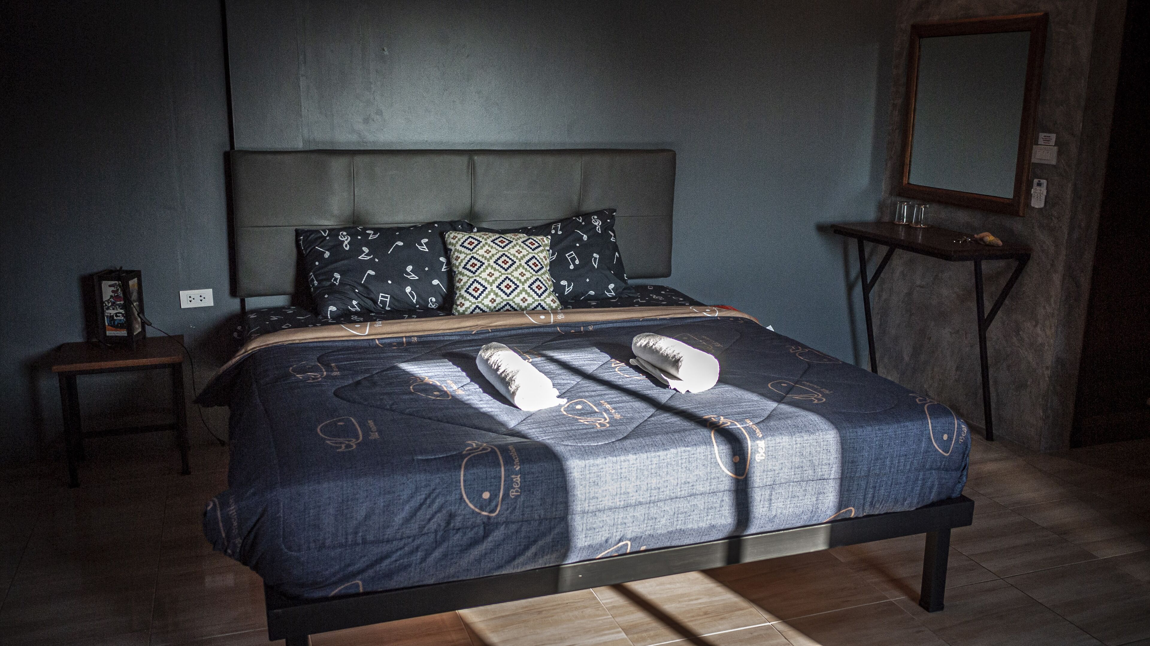 A bedroom with a bed, a bedside table, a nightstand, a mirror, and a wall socket.