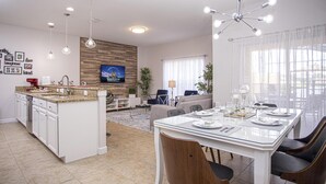 Una cuina moderna amb una illa central, una zona de menjador amb taula de vidre i un saló amb sofà i televisió.