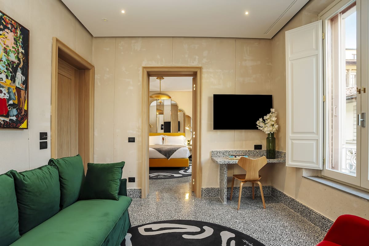 A hotel room with a green sofa, a small desk with a chair, a bed with a headboard, a flat-screen TV mounted on the wall, and a window with a view.