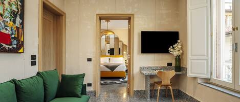 A hotel room with a green sofa, a small desk with a chair, a bed with a headboard, a flat-screen TV mounted on the wall, and a window with a view.
