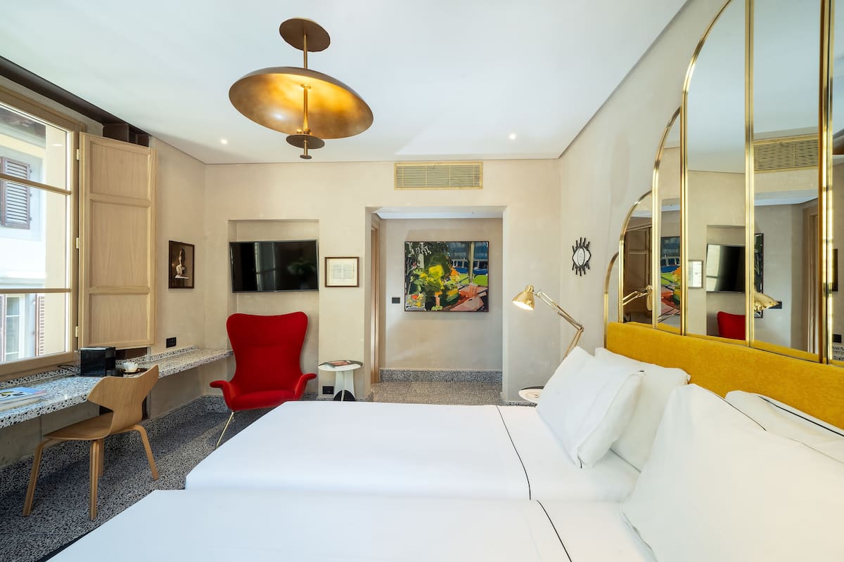 A hotel room with a bed, a desk, a red chair, a small table, a TV, and a large mirror.