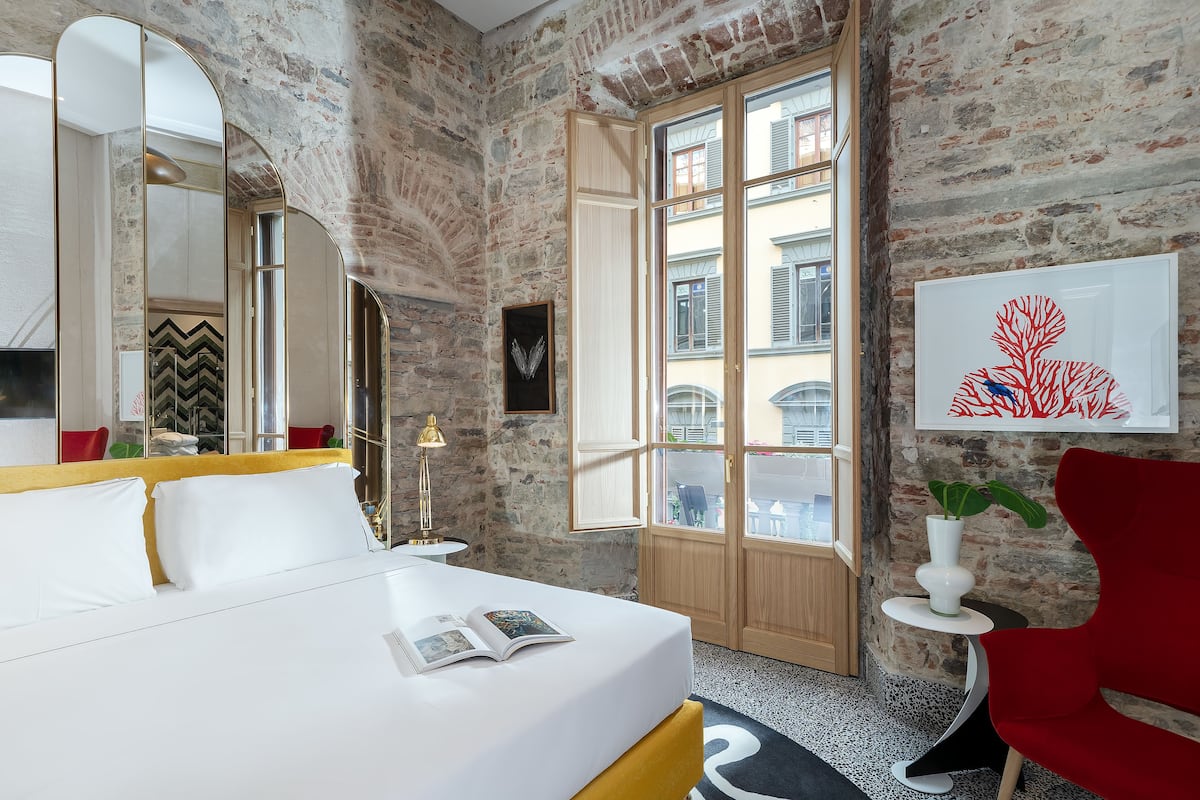A hotel room with a bed, a large mirror, a window with a view of buildings, and a red chair.