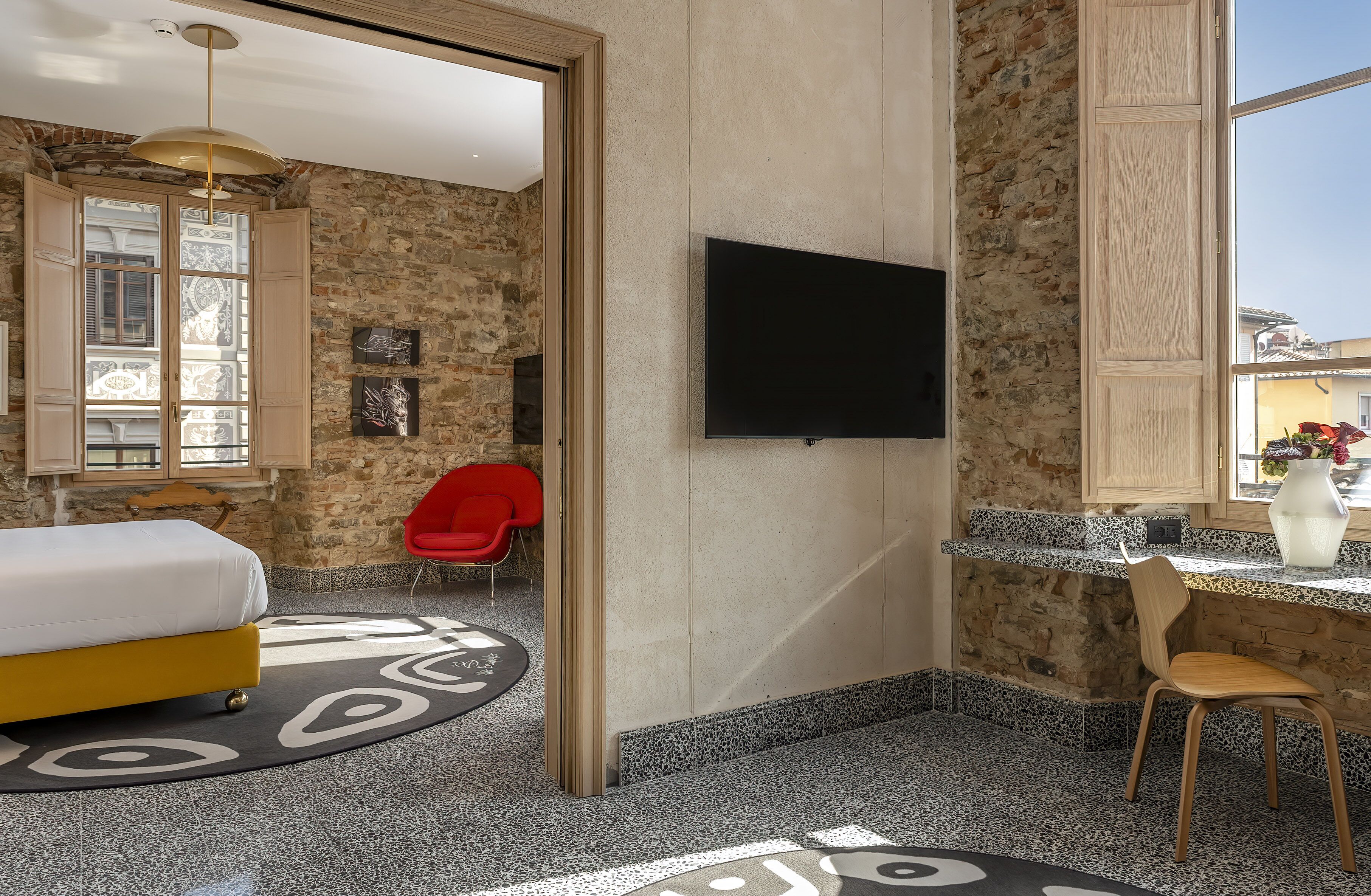 A modern hotel room with a bed, a TV, a red chair, and a stone wall.