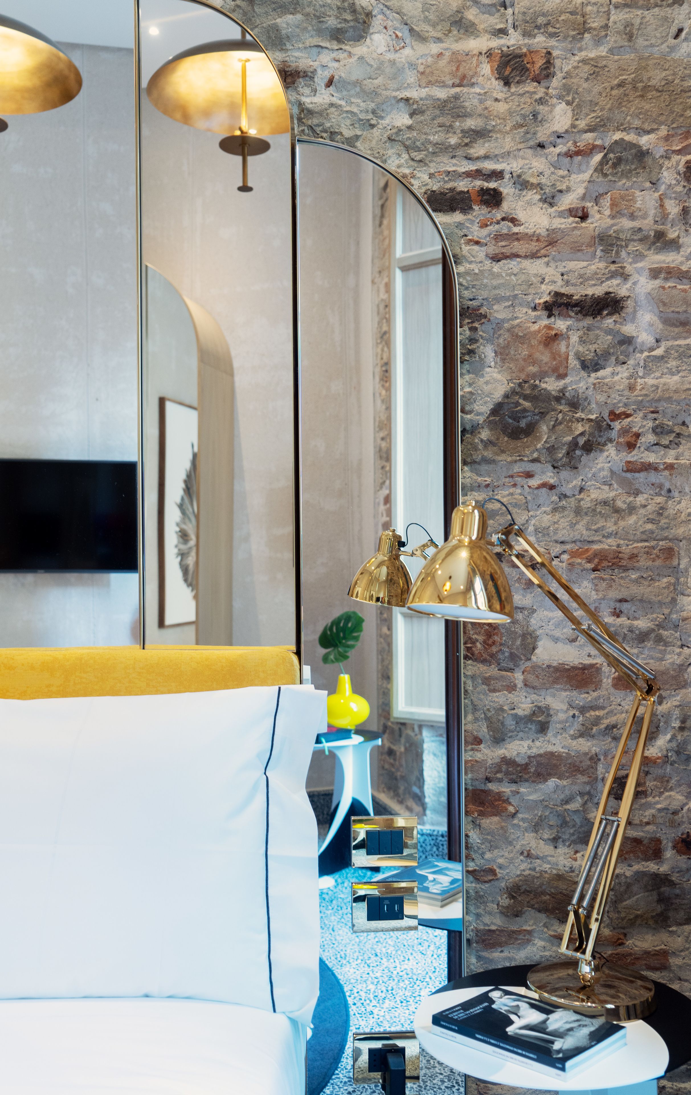 A modern bedroom with a stone wall, a bed with white bedding, a gold lamp, and a mirror reflecting the room.
