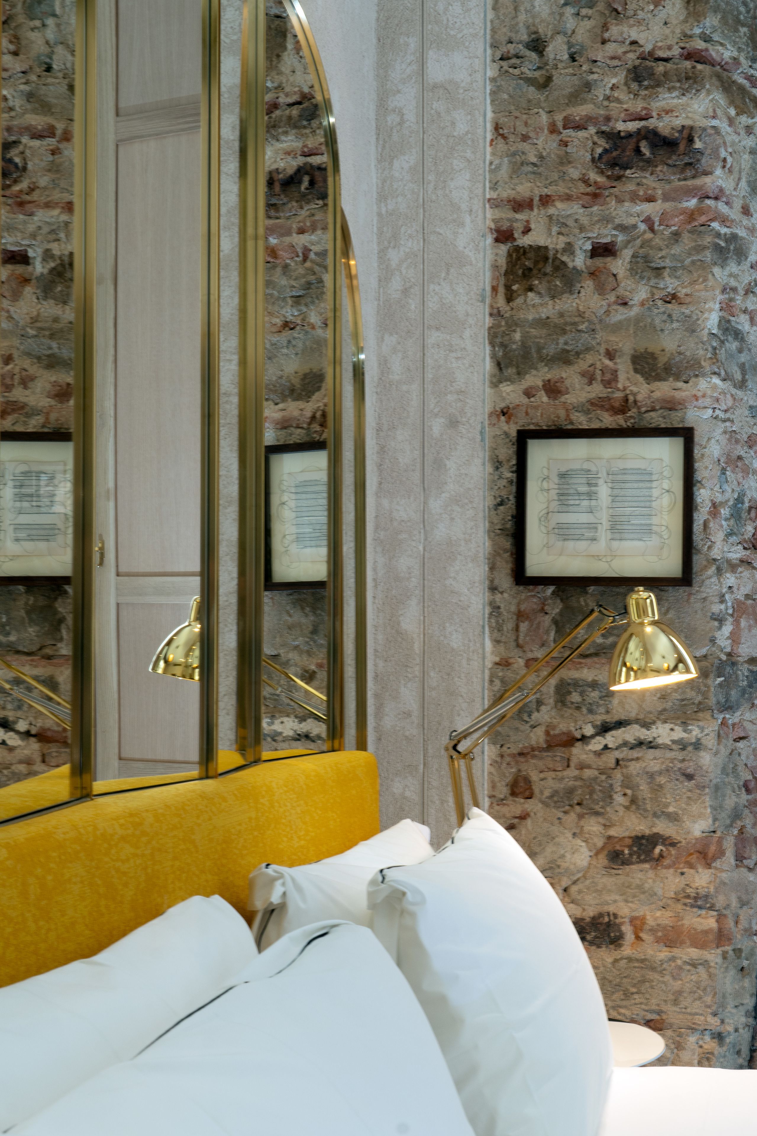 A bedroom with a brick wall, a large mirror, a gold lamp, and a yellow headboard.