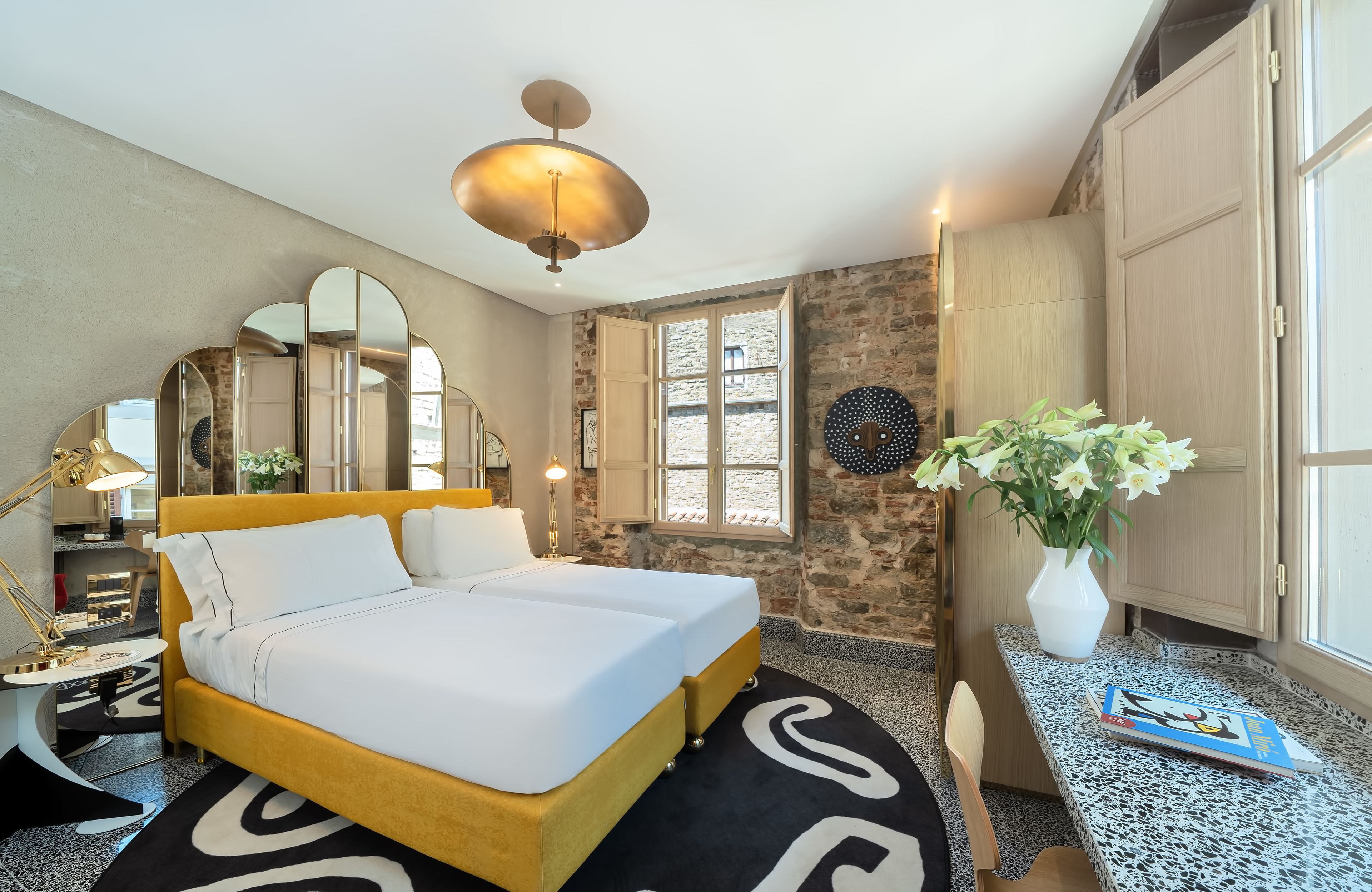 Ein Hotelzimmer mit einem Bett, einem Schreibtisch, einem Spiegel, einer Vase mit Blumen und einem Fenster mit Aussicht.