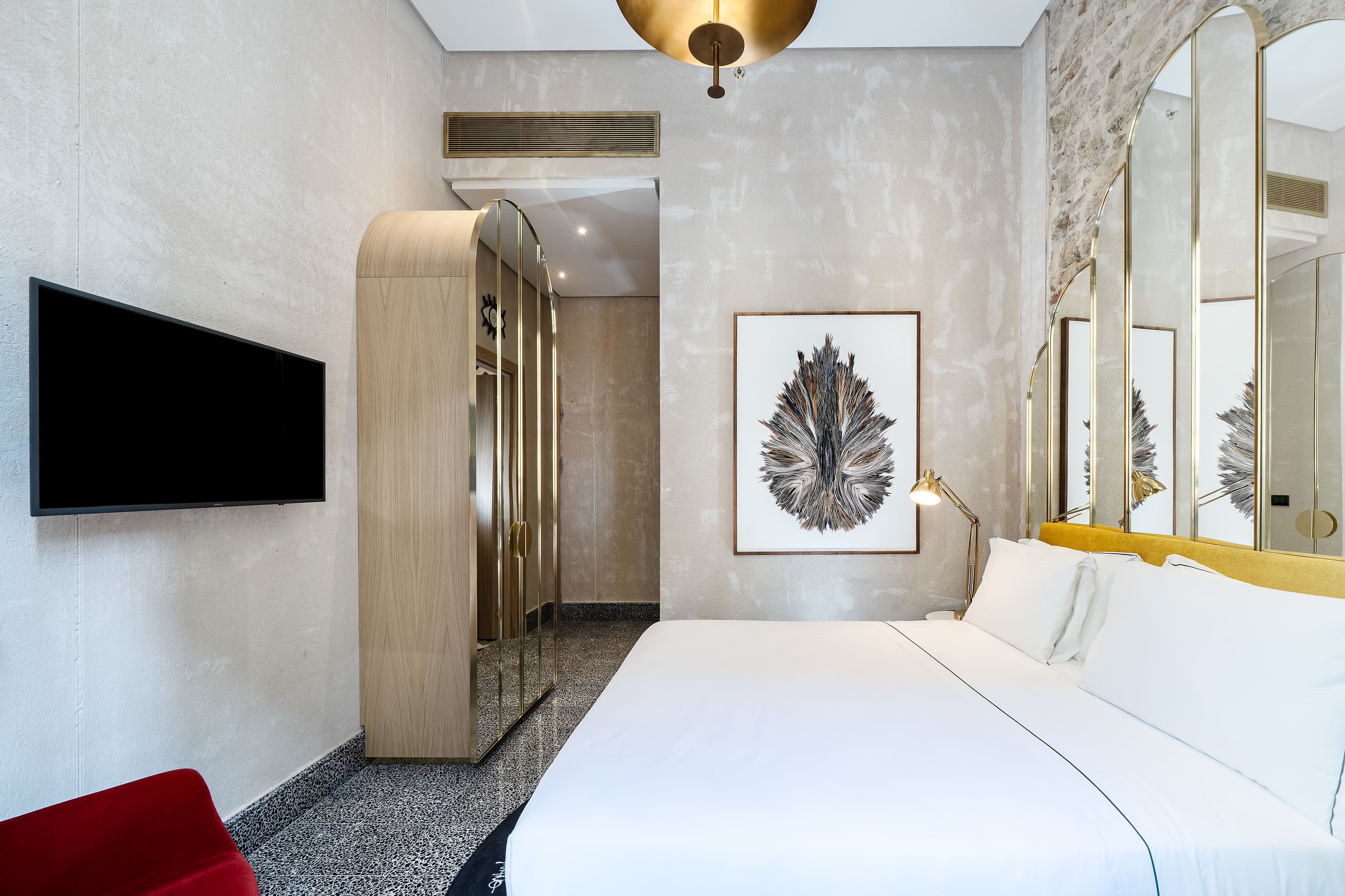 A modern hotel room with a large bed, a flat-screen TV mounted on the wall, a large mirror, and a unique wall art piece.