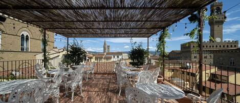 Un espace extérieur abrité, aménagé avec des chaises et des tables en métal blanc, offrant une vue sur une ville historique, avec un clocher imposant et une église.