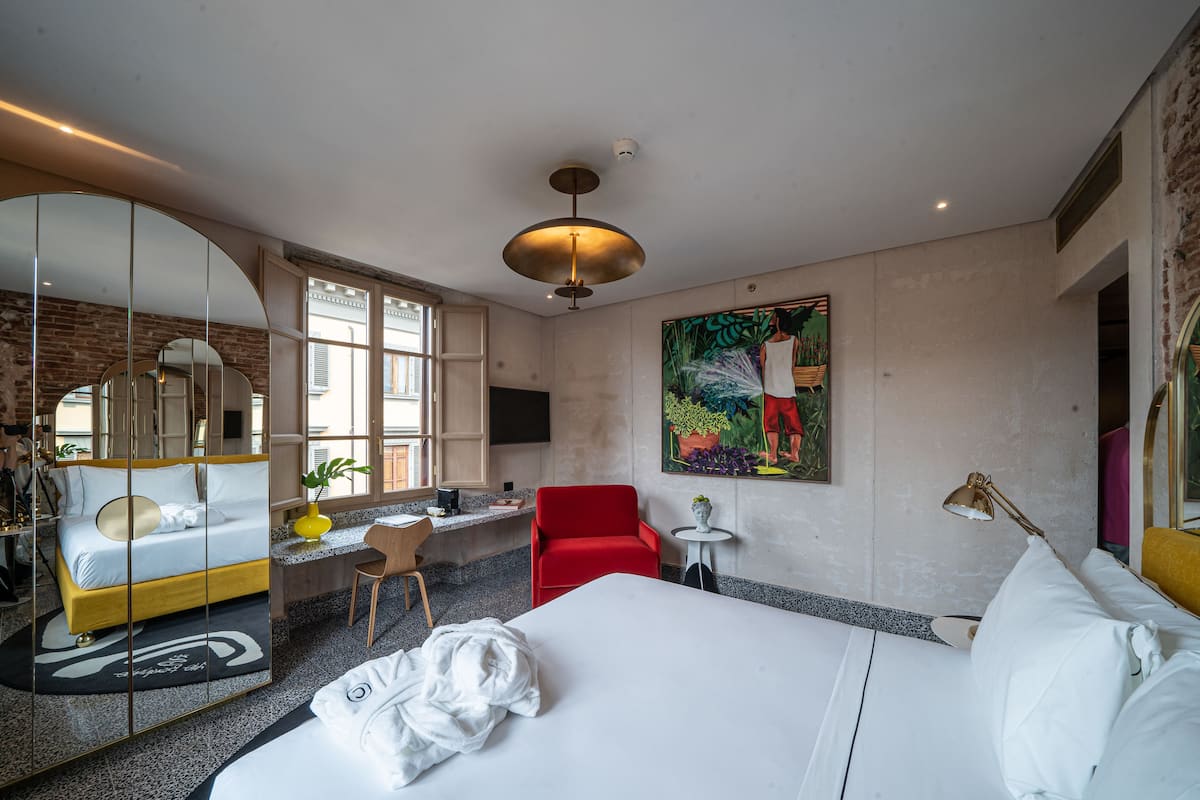 A hotel room with a large bed, a desk with a chair, a red armchair, a small table with a lamp, and a large painting on the wall.
