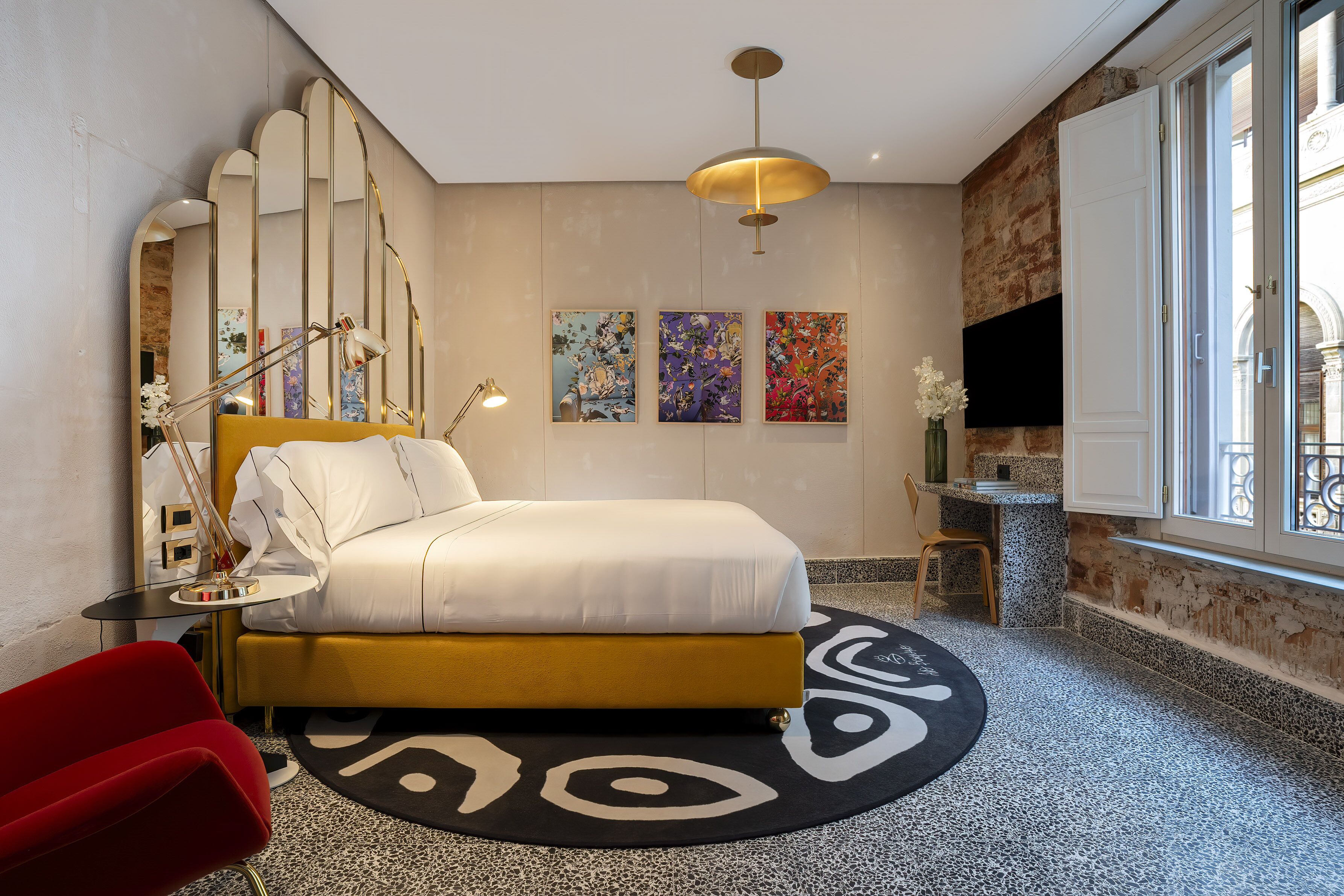 Una camera d'albergo moderna con un letto, una poltrona rossa, un tavolino con una lampada, una TV e opere d'arte alle pareti.