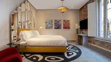 Una camera d'albergo moderna con un letto, una poltrona rossa, un tavolino con una lampada, una TV e opere d'arte alle pareti.