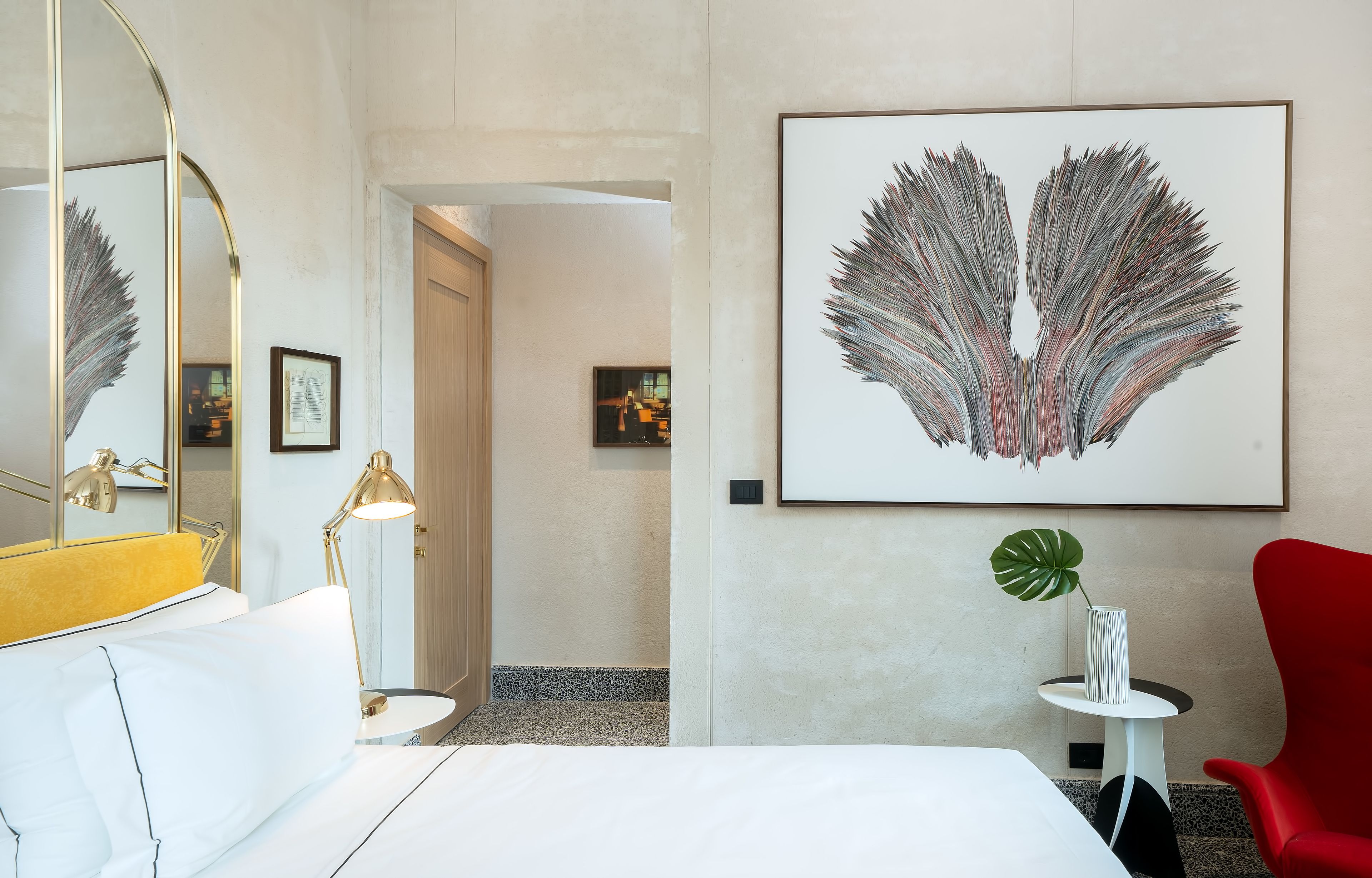 Une chambre d’hôtel avec un lit, une chaise rouge, une table ronde, un grand miroir et une œuvre d’art encadrée accrochée au mur.