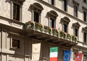 A building with ornate architectural details, a balcony with flowers, and flags including the Italian flag and the European Union flag.