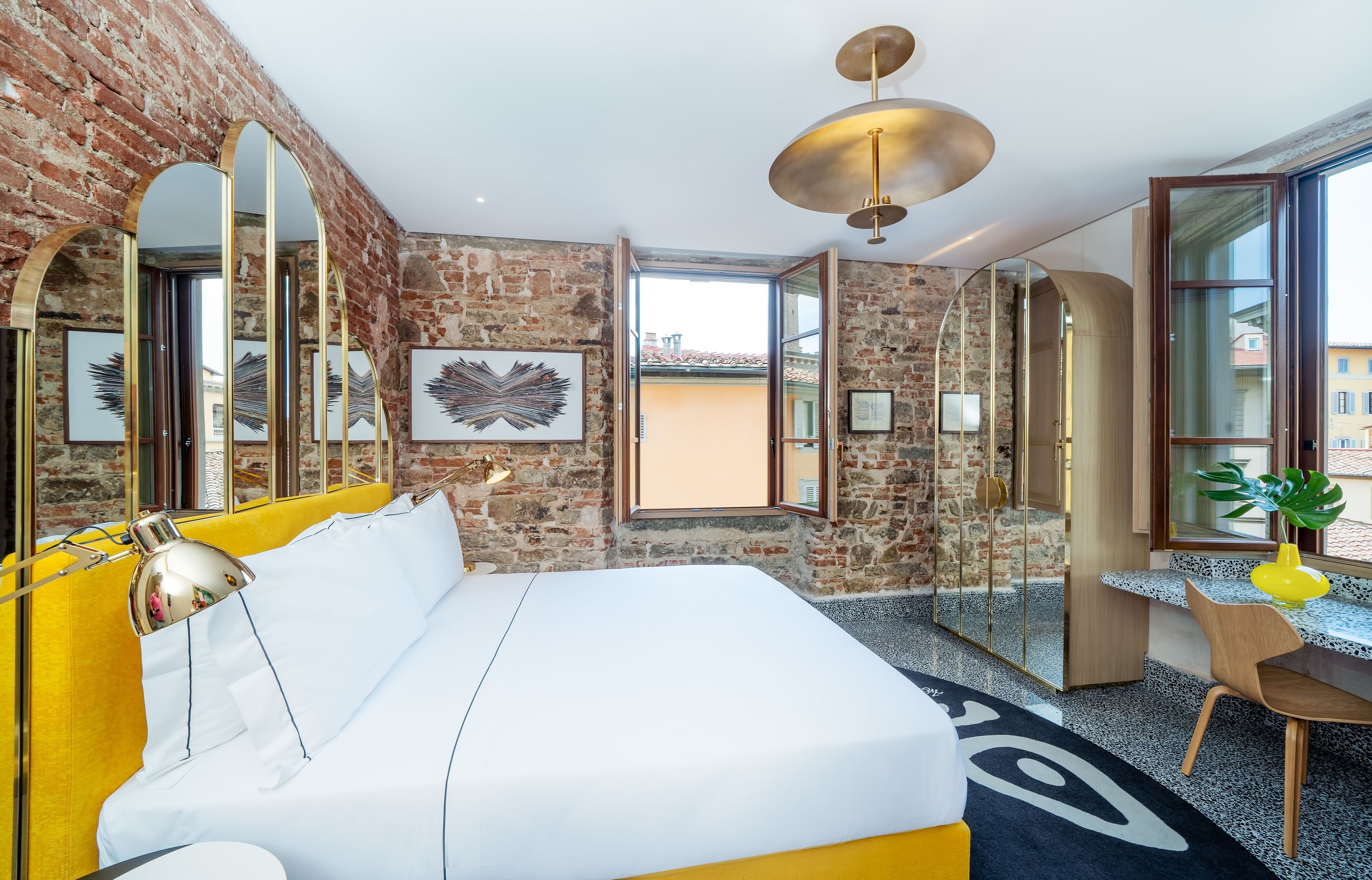 Ein Hotelzimmer mit Backsteinwand, einem großen Bett mit weißer Bettwäsche, einem Schreibtisch mit Stuhl und einem großen Fenster.