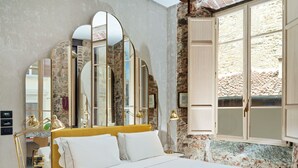 A bedroom with a bed, a desk, and a large window with stone walls.