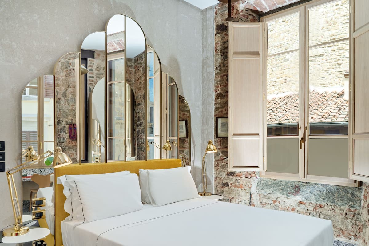 A hotel room with a bed, a desk, a chair, a mirror, and a window with a stone wall.