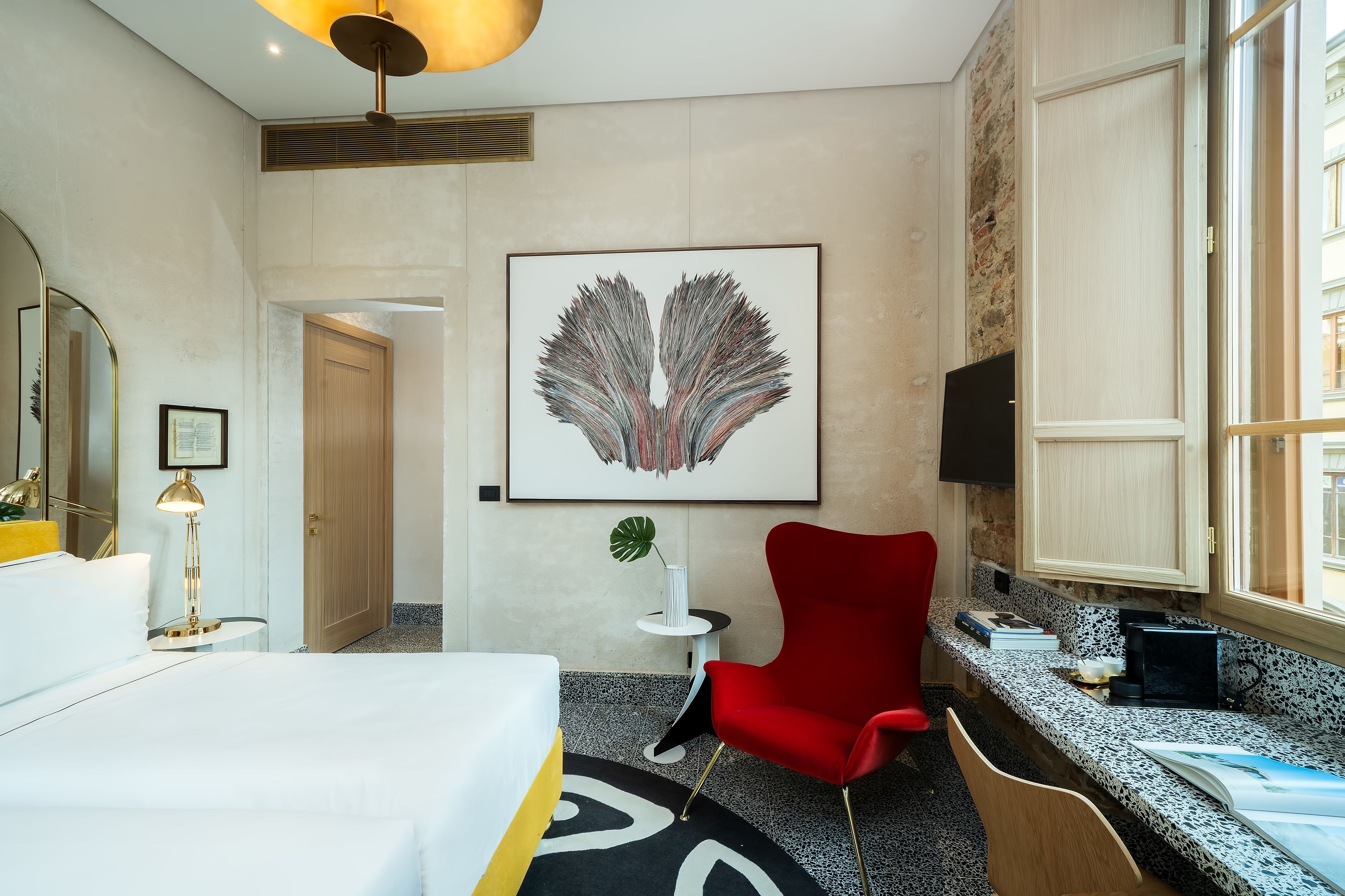 A hotel room with a bed, a red chair, a desk with a computer, a large artwork on the wall, and a window with a view.
