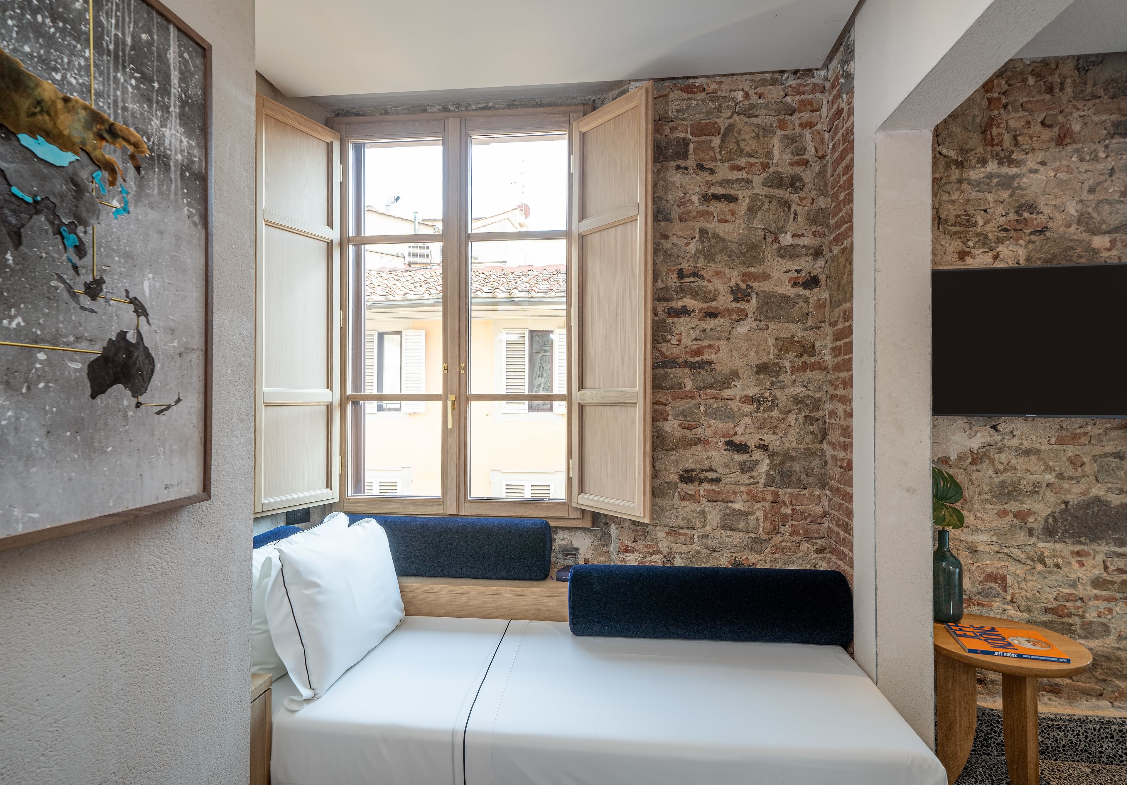 Una habitación con una cama, una ventana con contraventanas abiertas, un muro de ladrillo y un televisor montado.