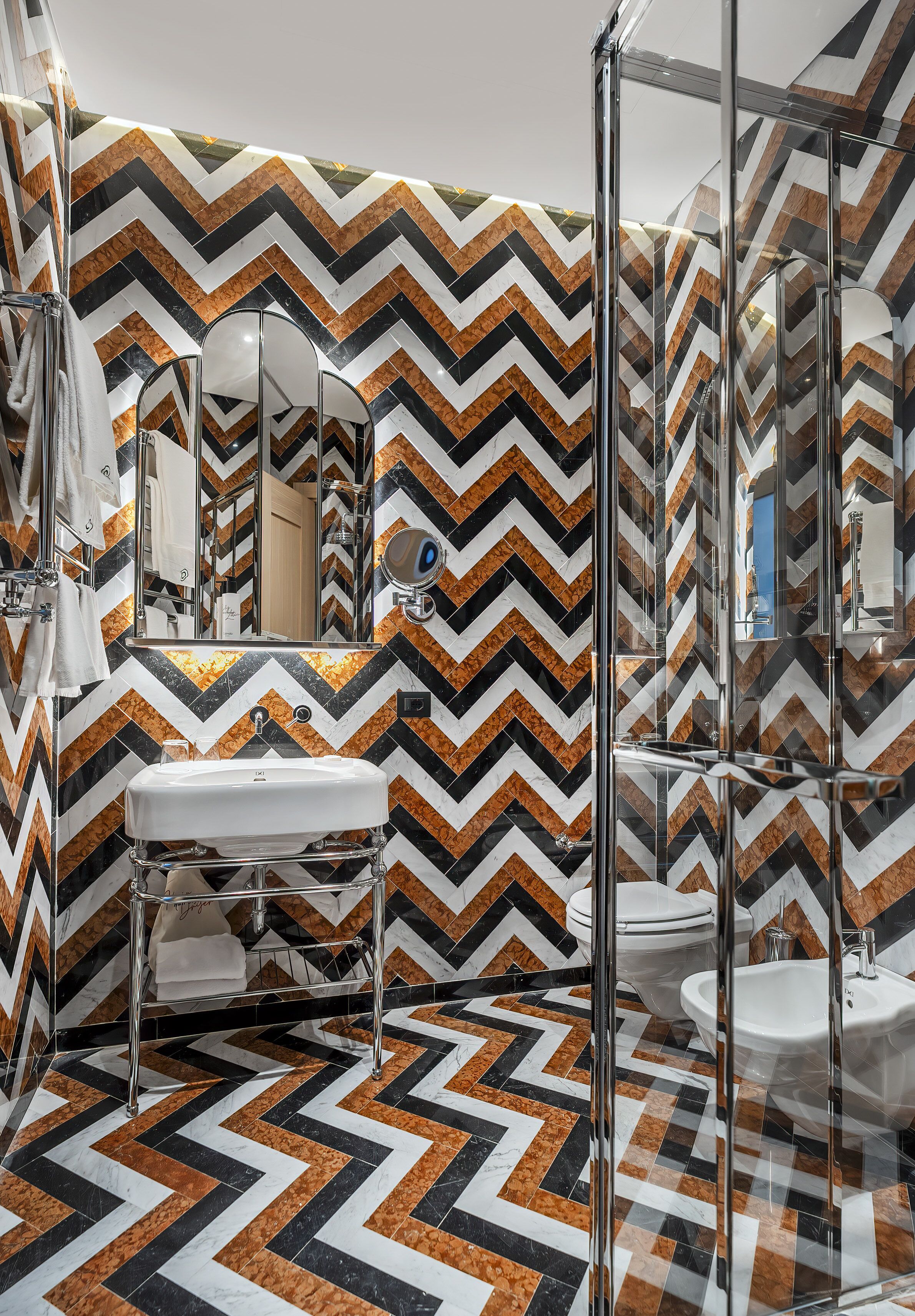 Une salle de bain moderne avec un mur recouvert de carreaux disposés en motif chevrons, une douche en verre et un lavabo blanc.