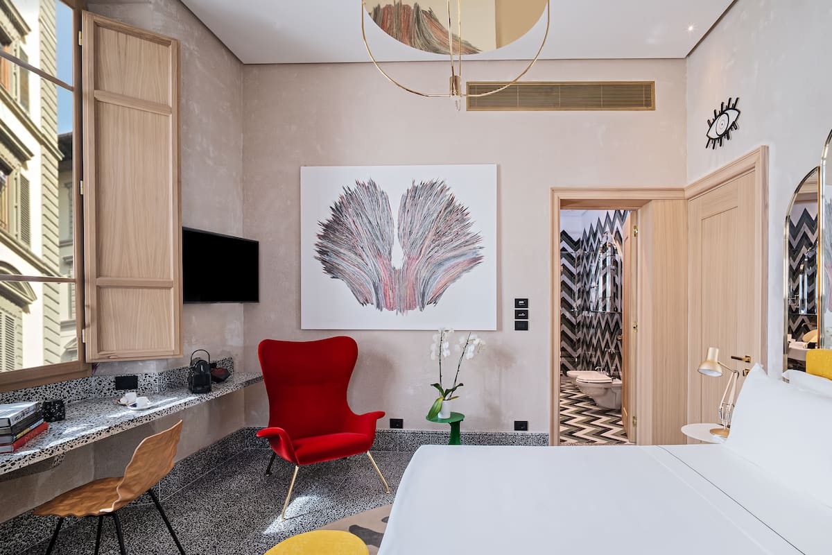 A modern hotel room with a large window, a red chair, a desk with a computer, a white bed, and a large abstract painting on the wall.