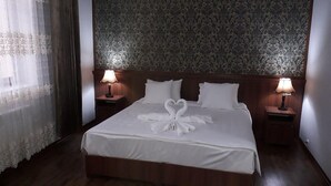A neatly made bed with white linens and a heart-shaped towel arrangement, two bedside lamps, and a patterned wallpaper. - Hotel Erkin Palace (Khiva)
