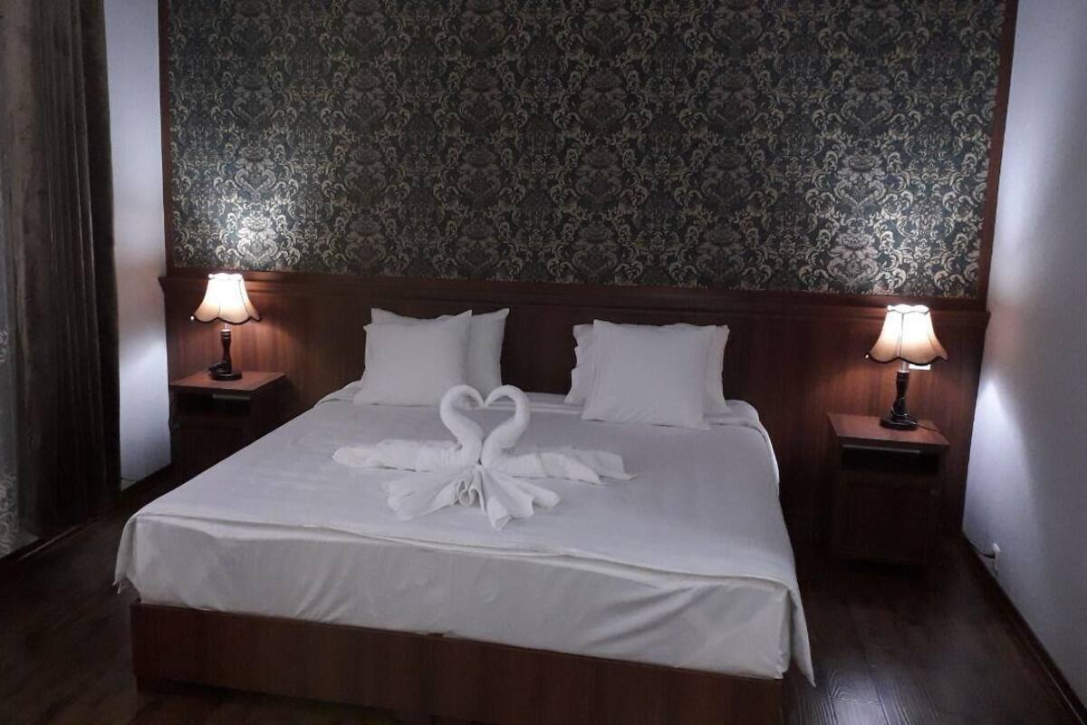 Una cama bien hecha con sábanas blancas y una decoración de toallas en forma de corazón, dos lámparas de noche y un papel tapiz estampado.