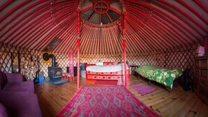 A traditional yurt interior with a central bed, a sofa, a fireplace, and a wooden floor. - Big Red Yurt at Cabot Shores Wilderness Resort (Englishtown)