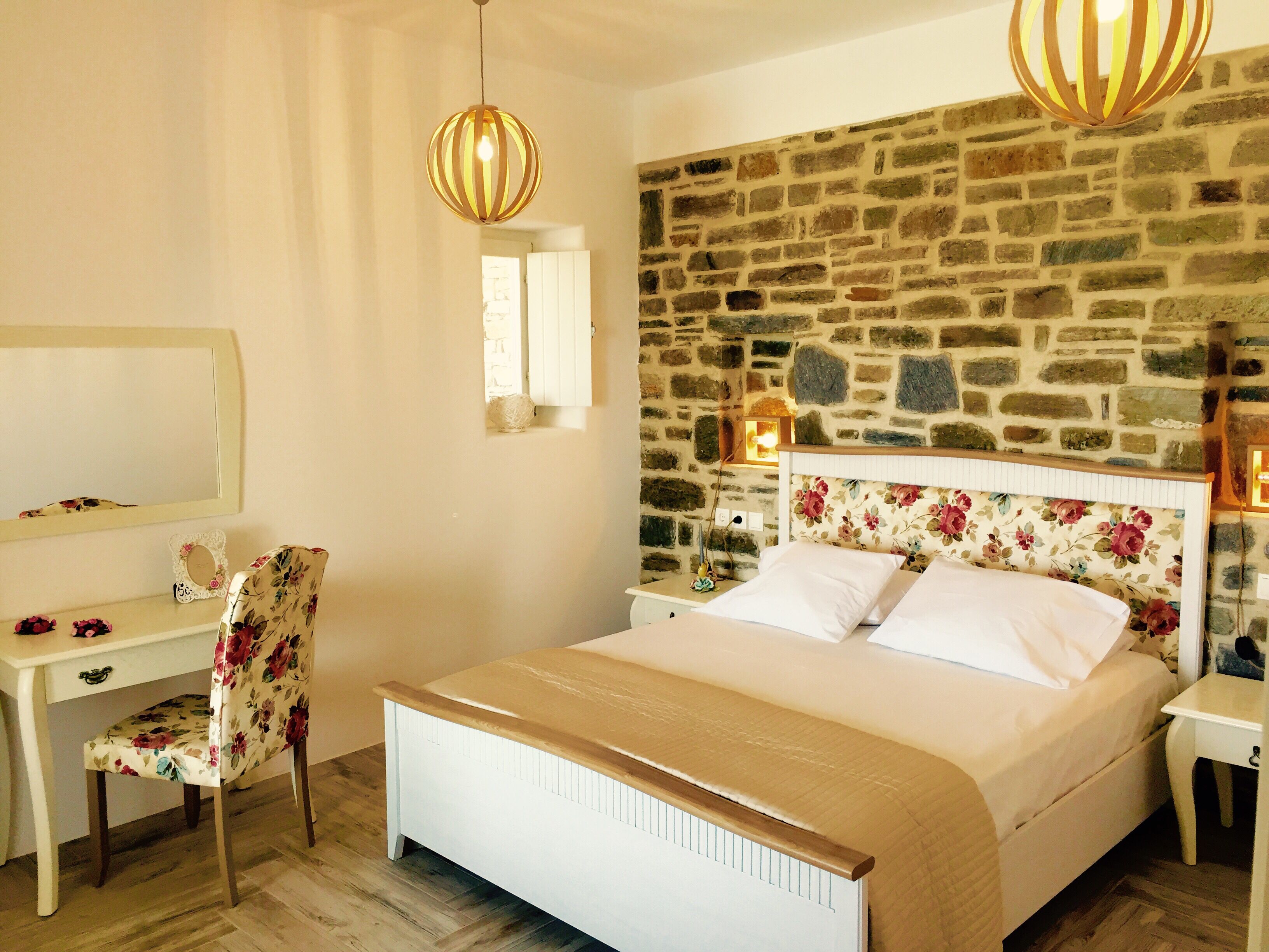 A bedroom with a stone wall, a bed with floral headboard, a desk with a chair, and two hanging lamps.