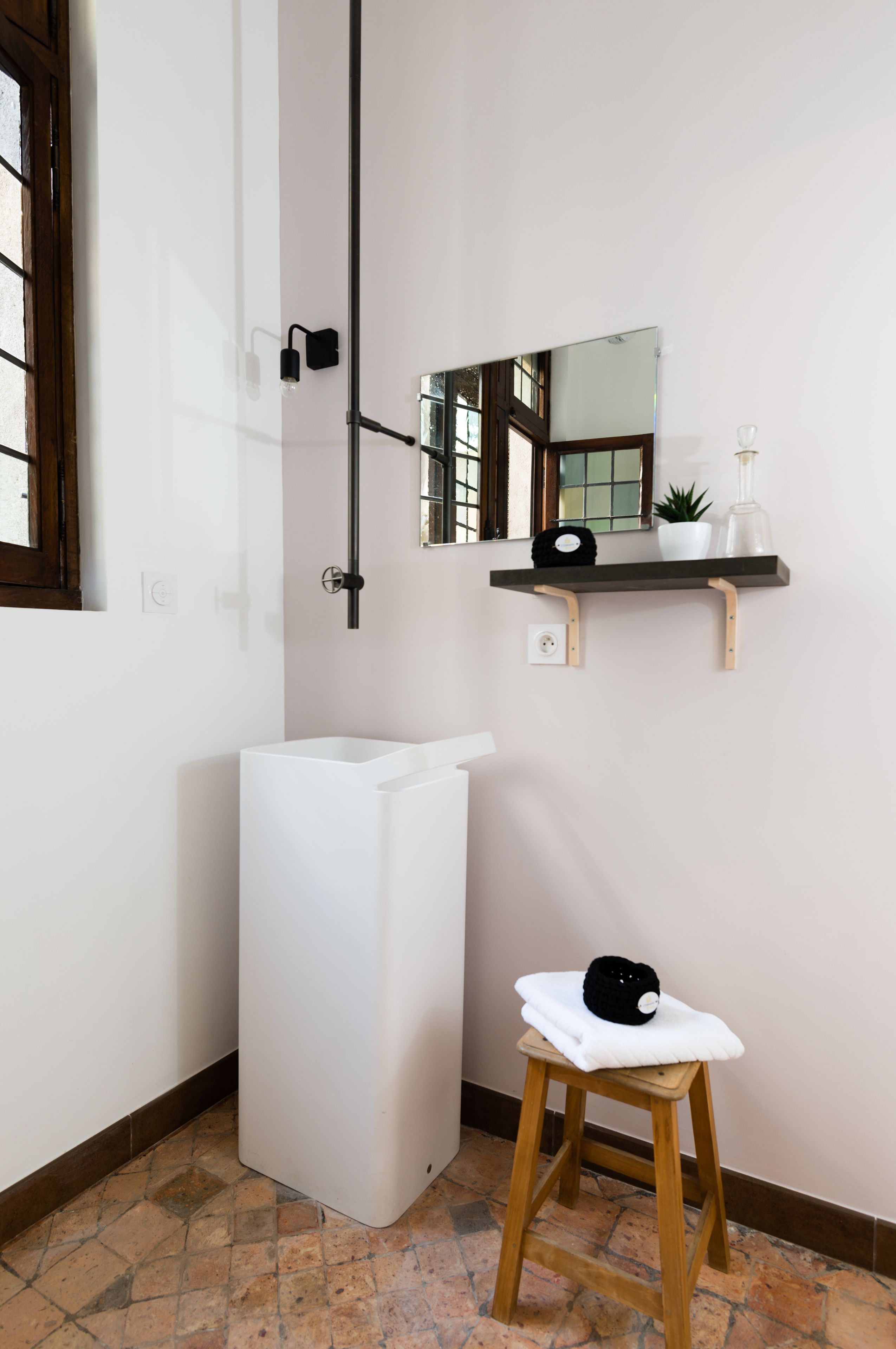 Un baño con un lavamanos blanco, un taburete de madera, un espejo y una estantería con una planta y una botella.