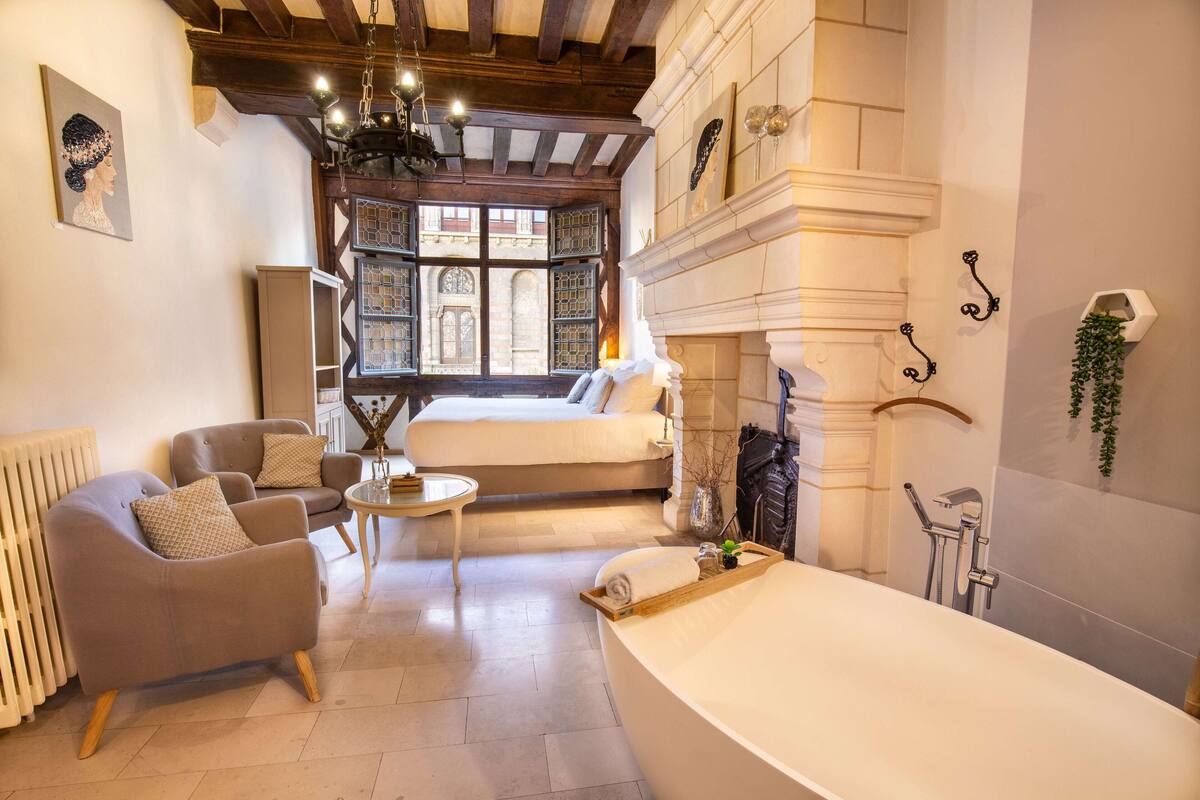 A spacious room with a large bed, a sitting area with a chair and table, a fireplace, and a view through a window with a decorative door.