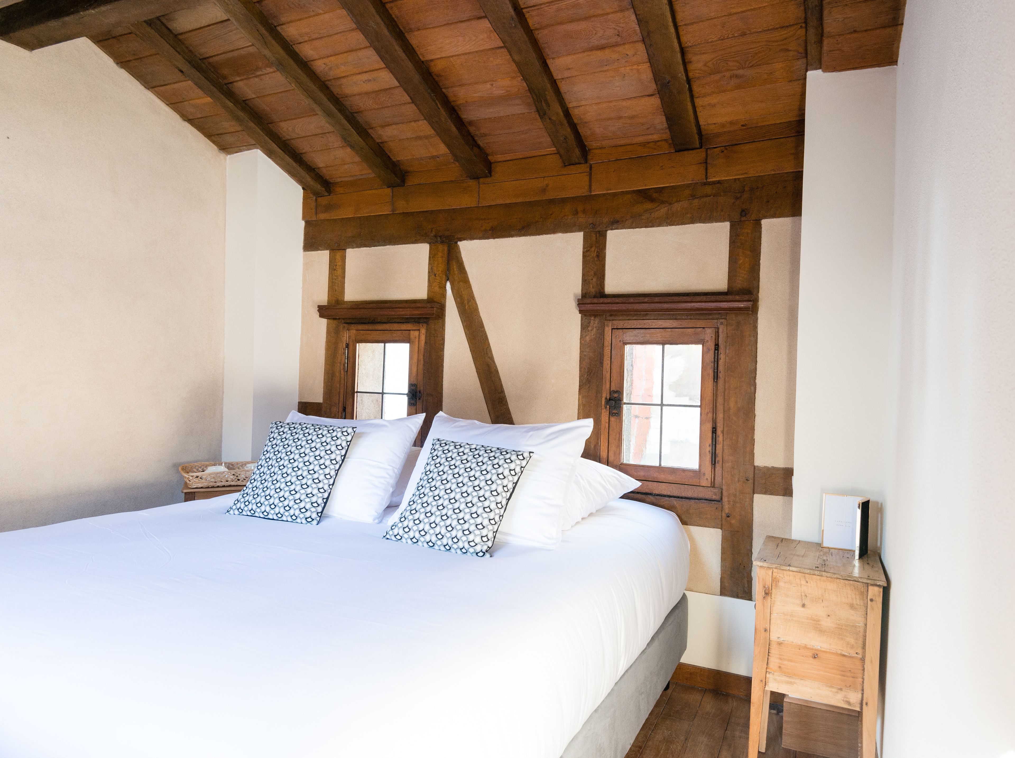 Een slaapkamer met een groot bed, houten balken in het plafond, twee ramen en een klein houten nachtkastje.