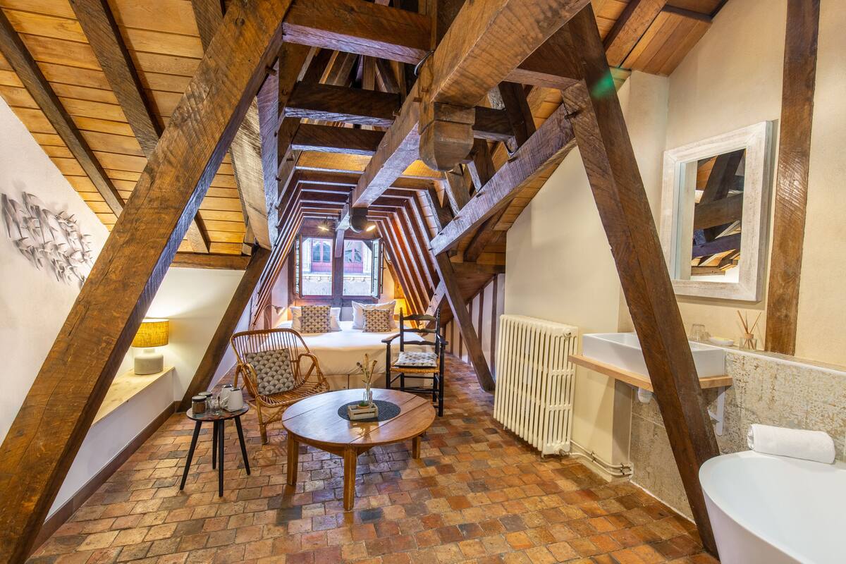 A cozy attic bedroom with exposed wooden beams, a brick floor, a round wooden table, two cushioned chairs, a bed with a headboard, and a window with a view.