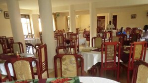 A dining area with multiple tables and chairs, large pillars, and a person in the background. - Margarita Palace Hotel (Lira)