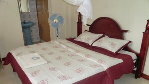A double bed with a floral bedspread and a red border, a bedside table, a wall-mounted fan, and a wooden headboard. - Margarita Palace Hotel (Lira)